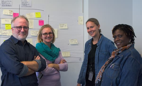 Four people standing and smiling in front of a white board covered with sticky notes