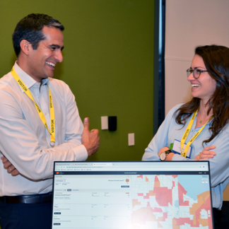 Man and woman standing behind a computer monitor facing each other and smiling.